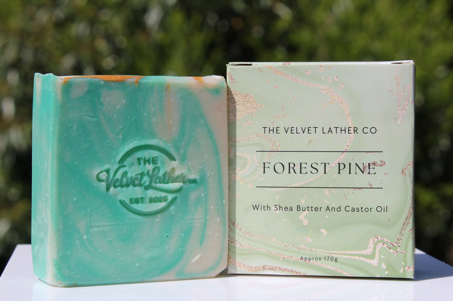 Bar of soap and packaging of 'The Velvet Lather Co' Forest Pine soap on a white surface with a blurred green background.