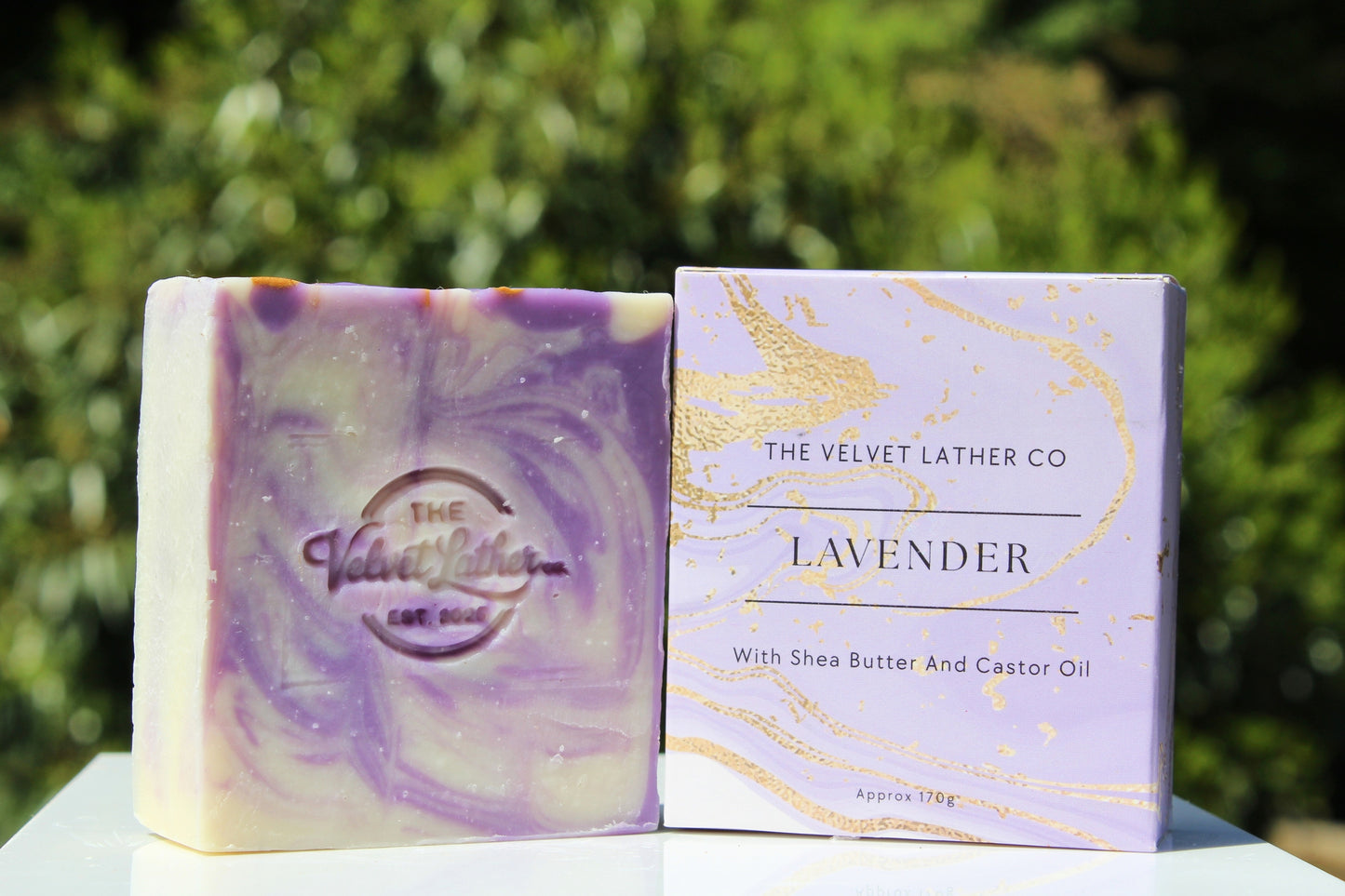 Lavender soap bar and packaging with 'The Velvet Lather Co' branding against a green outdoor background.