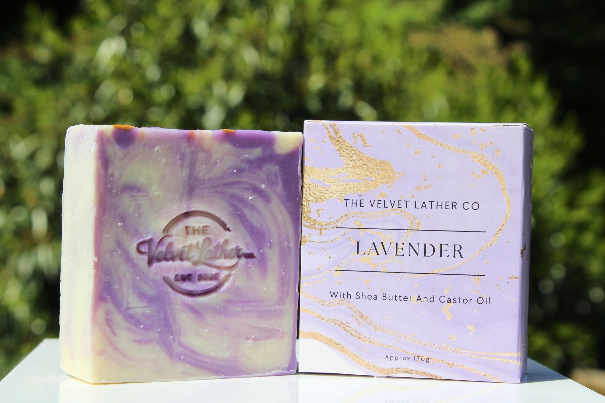Lavender soap bar and packaging with 'The Velvet Lather Co' branding against a green outdoor background.