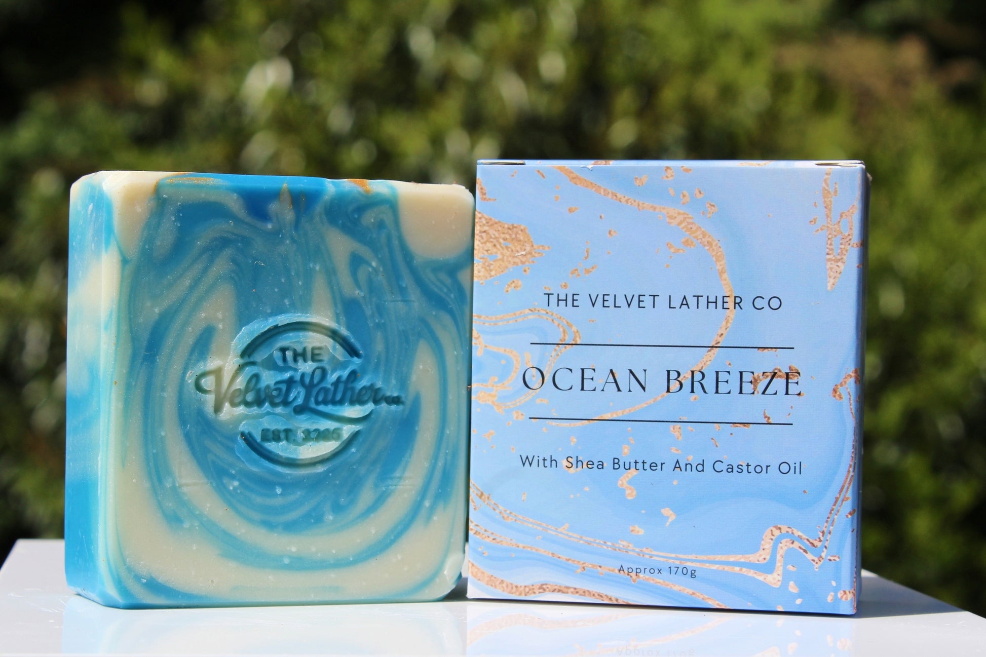 Bar of soap with marbled design and packaging labeled 'The Velvet Lather Co. Ocean Breeze' on a natural background.