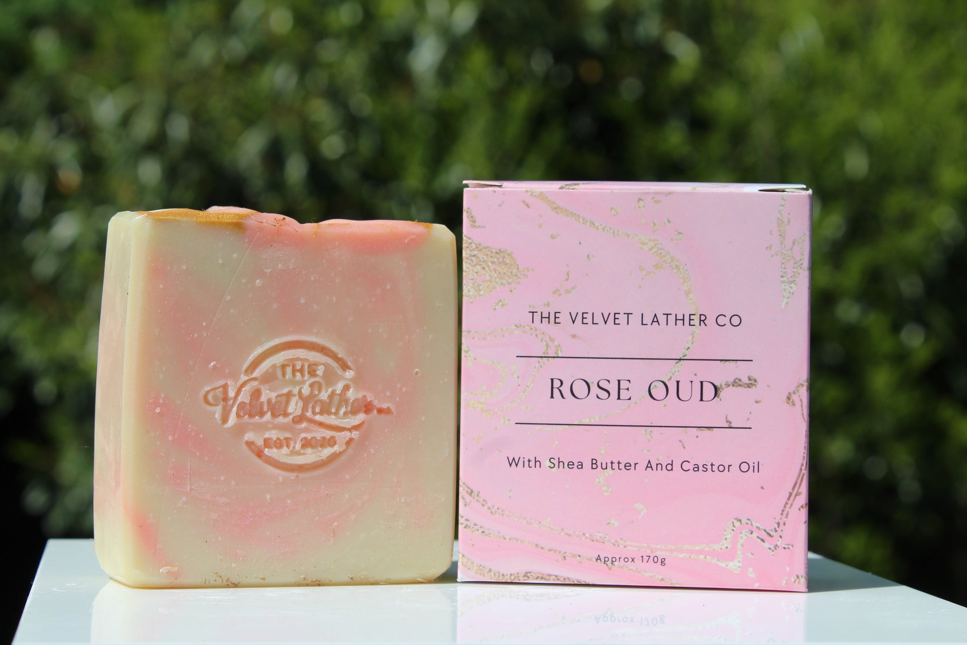 Bar of soap and packaging box from The Velvet Lather Co on a reflective surface with greenery in the background